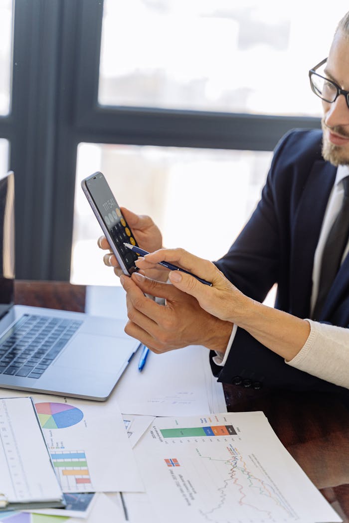 Home Business professionals reviewing financial reports on a smartphone at a desk with papers and a laptop in an office setting.